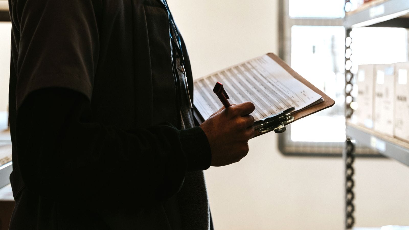 A person in a dark jacket and lanyard holding a clipboard of ruled forms with a pen, standing beside stockroom shelving with a bright window behind